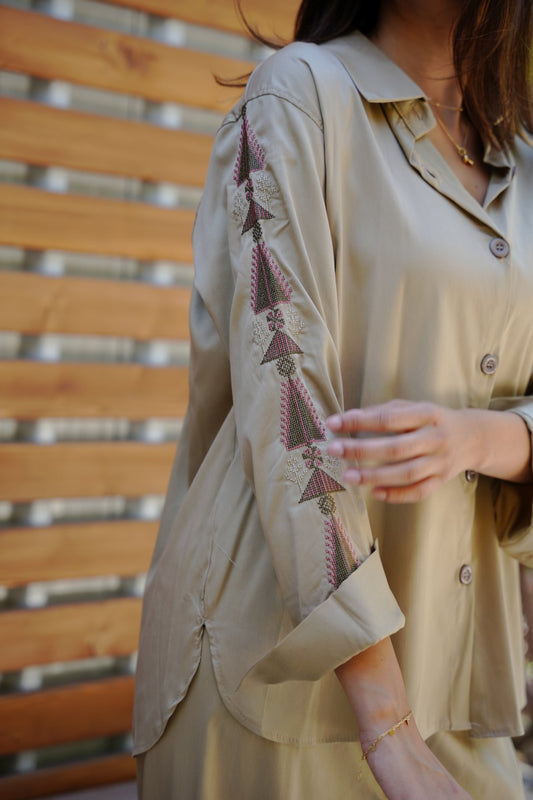 Person wearing a beige coat with intricate tatreez embroidery patterns, sitting on a wooden bench.